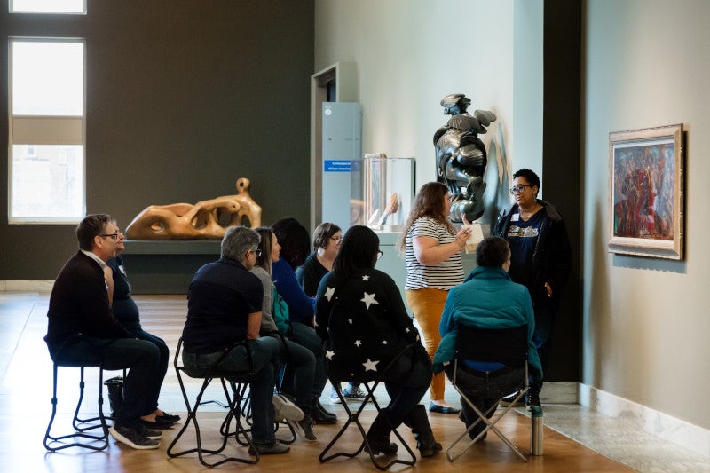 An adult group sits on gallery chairs while listening to a DIA Gallery Teacher