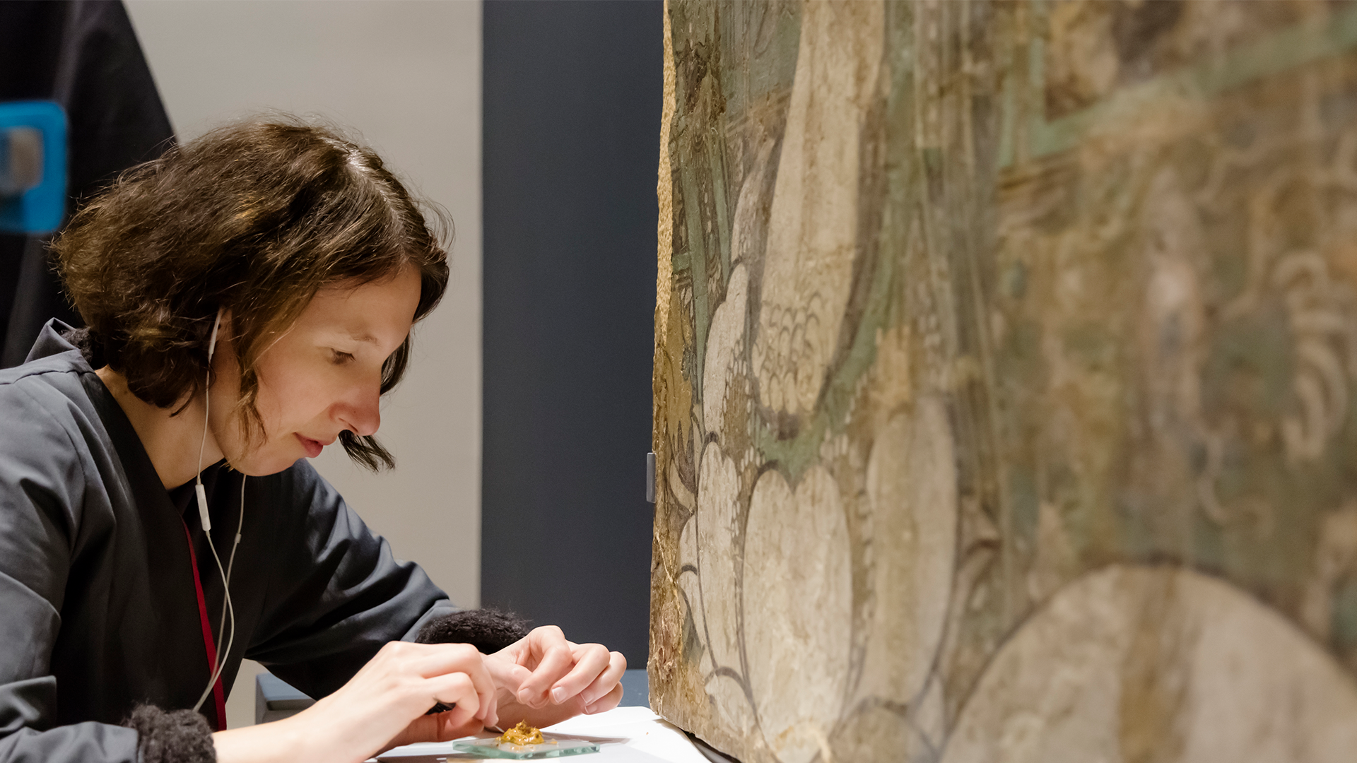 A conservator wearing white headphones mixes up a small mound of gold paint next to a large painted work