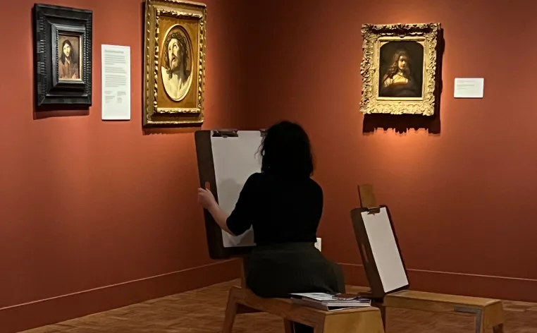 A visitor in all black sits on an easel bench drawing in the Dutch galleries