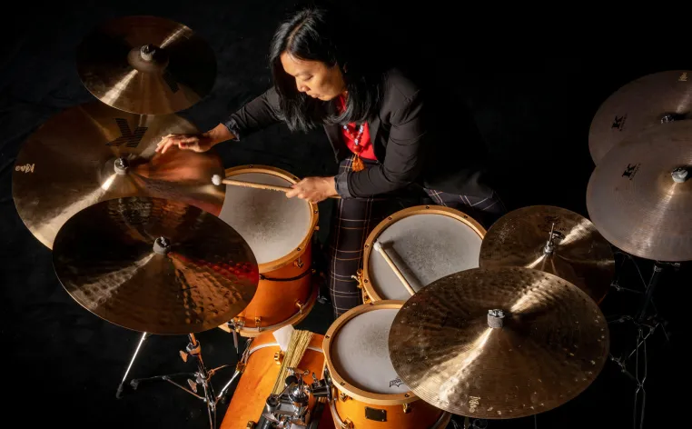 Suzie Ibarra, wearing a suit with plaid pants, sits and plays a set of drums