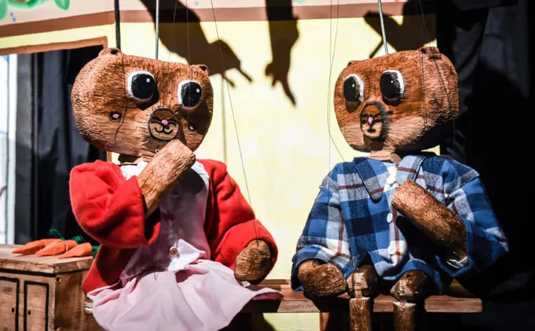 Two wooden puppet bears sat next to each other on a wooden bench as if in active conversation.
