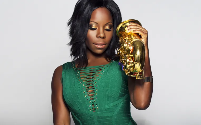 A Black woman poses in an emerald green dress while holding a saxophone on her left shoulder.