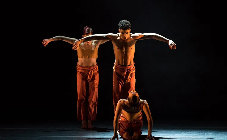 Three dancers on stage, two standing with their arms up and one sitting on the ground, looking backward towards the camera