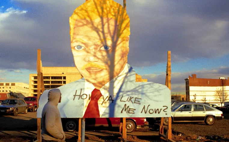 A person in a gray hoodie with the hood up stands shadowed in front of a large cutout sign of a head.