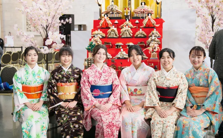 Six women, in traditional Japanese garments, sit in a row and smile at the camera.