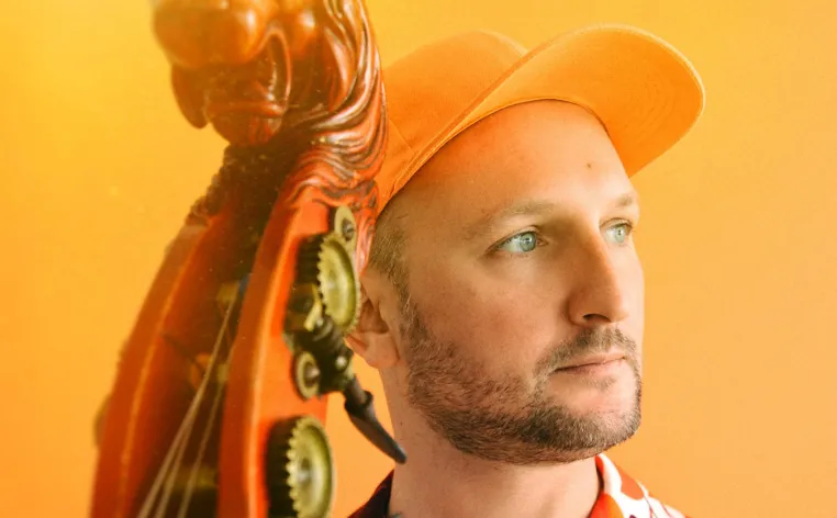Florent Ghys pictured with his bass in a orange baseball cap in front of an orange background.