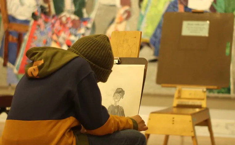 A person in a hoodie and beanie sits on an easel stool drawing in the *Contemporary galleries.