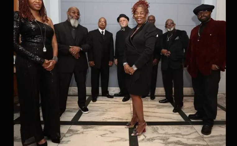 A large group of musicians stand together, dressed in black