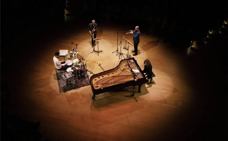 Two performers, a drum kit, and a piano lit on a dark stage, pictured from above.