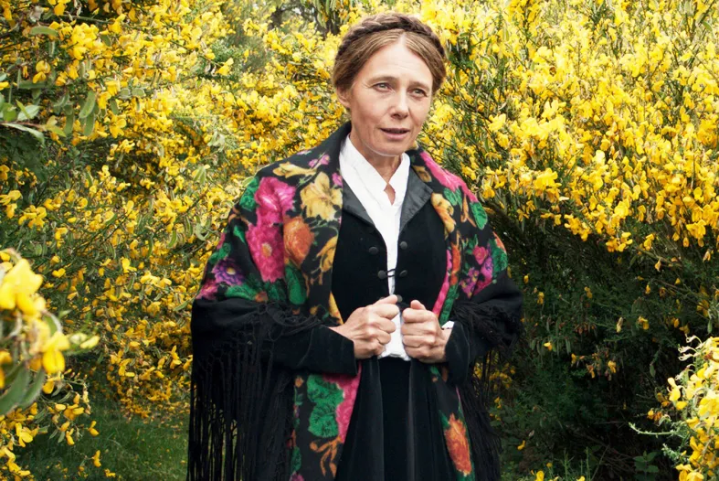 A woman in a black robe with a bright floral shawl stands amongst trees.