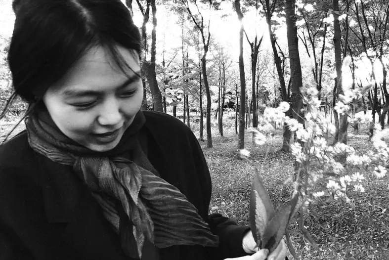 A woman bundled in a coat and scarf holds flowers picked in a forest, pictured behind her.