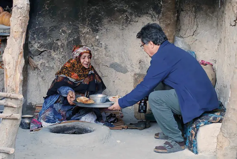 A man and a woman in a natural dwelling handing each other food over an in-ground fire pit