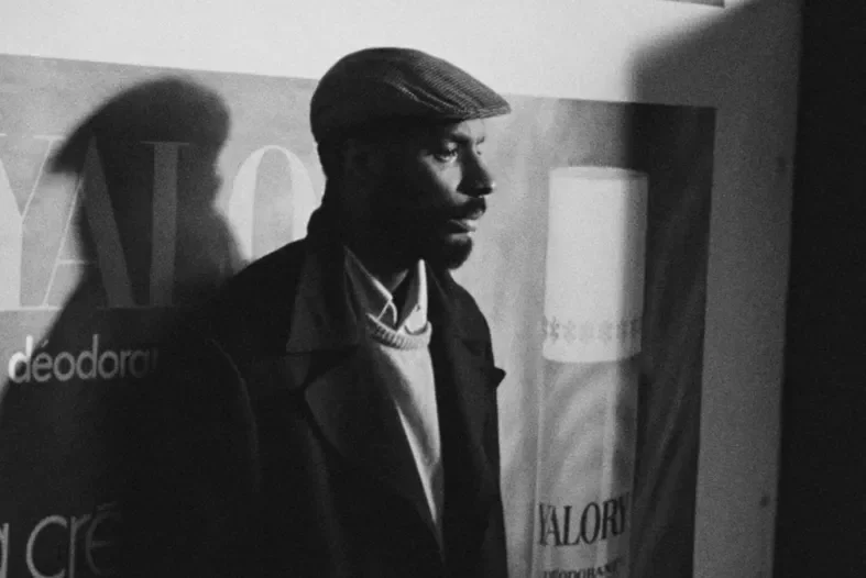 Black and white image of a man standing forlornly in front of a deodorant ad in a sweater, coat, and flat cap.