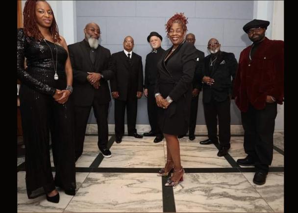 A large group of musicians stand together, dressed in black