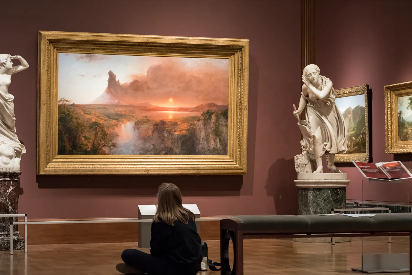 A visitor sitting on the floor next to a bench in front of Frederick Church's "Cotopaxi"