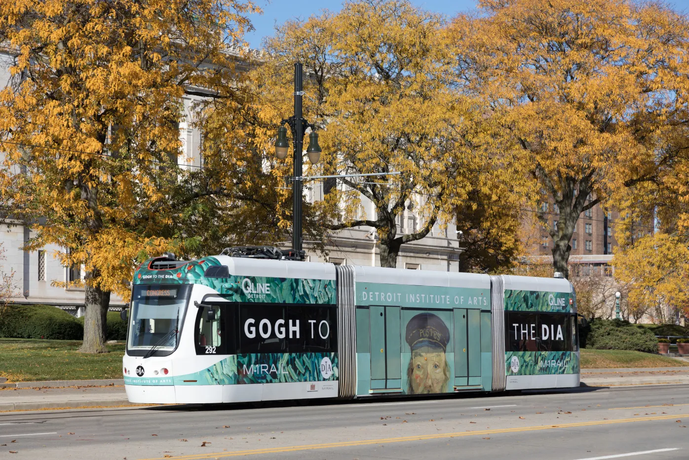 The Detroit QLine pictured amongst fall trees with a wrap featuring the DIA&#039;s iconic &quot;Postman&quot; by Van Gogh and the words Gogh to the DIA