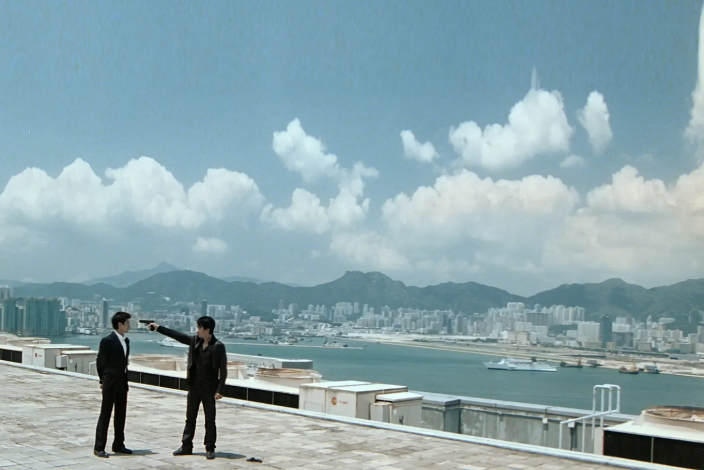 Two figures stand atop a high-rise roof with the city, a river, mountains and copious clouds behind them.