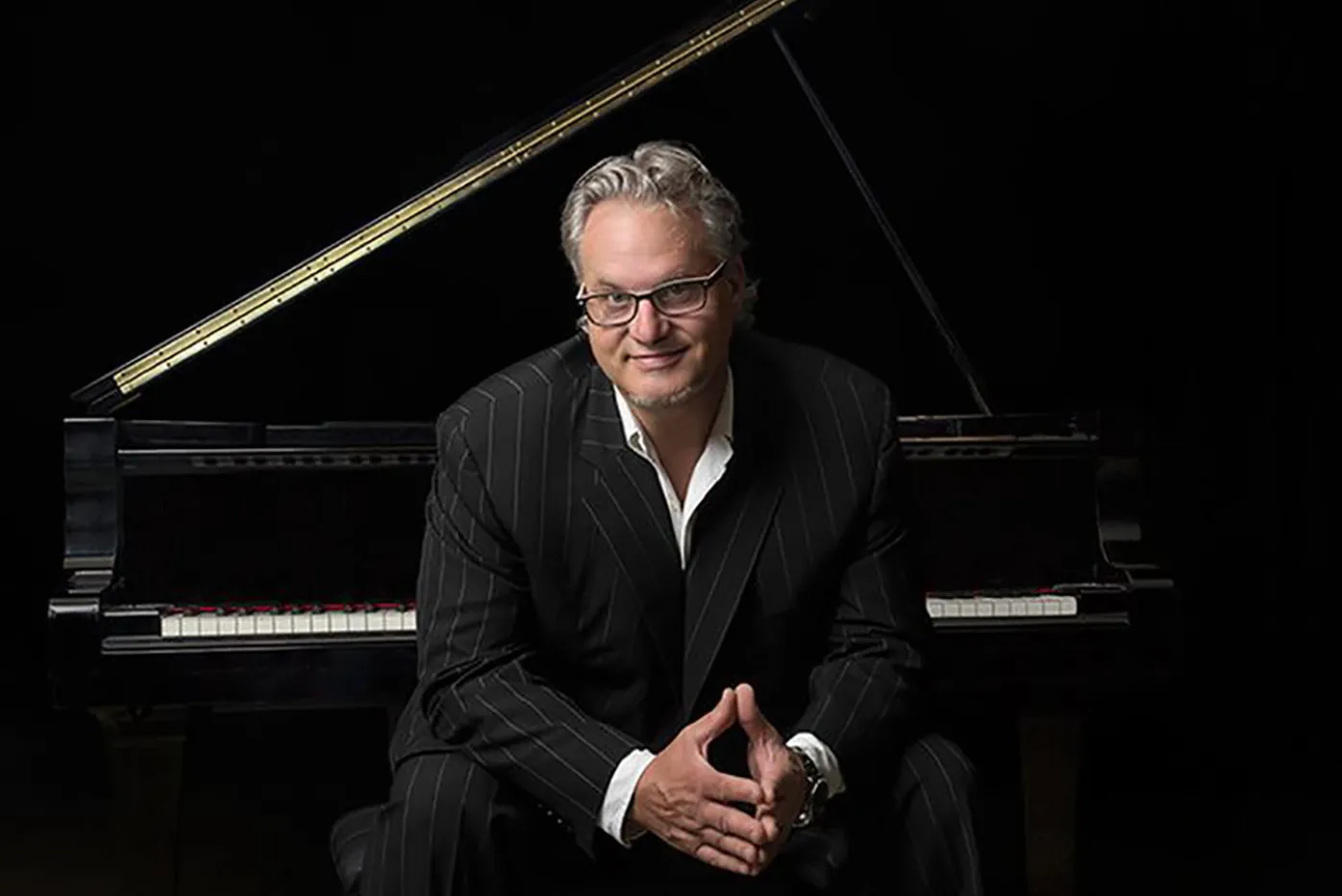 Mr. B in a suit with an open collar, seated in front of a grand piano.