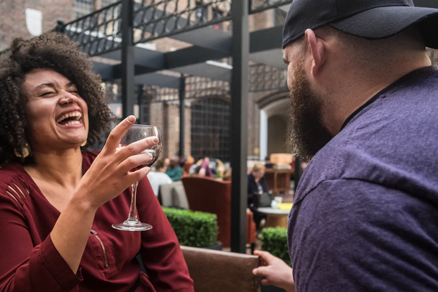 A woman holds a glass of white wine as she laughs while chatting to a man in a backwards baseball cap.