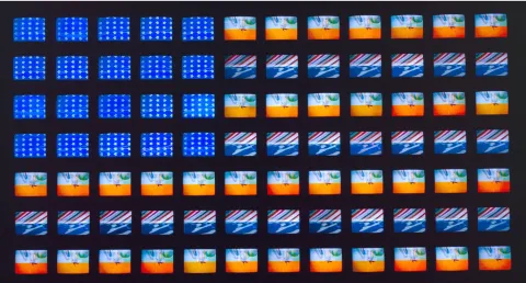 Image of Video Flag XGrid of small video screens arranged like a flag, displaying shifting patterns of blue fields with white stars and horizontal bands of red, white, and abstract imagery. The screens vary slightly in color and motion, creating a flickering, electronic interpretation of the American flag in Video Flag X by Nam June Paik.
