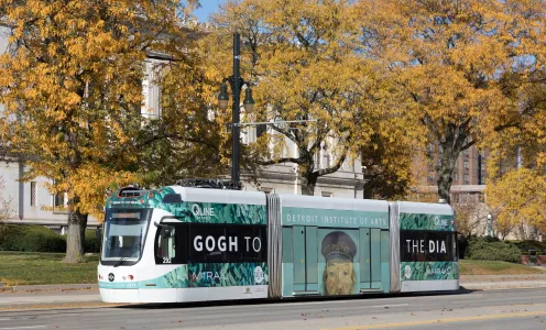 The Detroit QLine pictured amongst fall trees with a wrap featuring the DIA's iconic "Postman" by Van Gogh and the words Gogh to the DIA