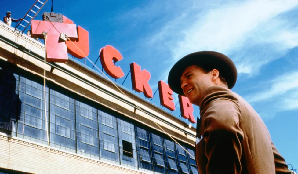 A man with a wide brimmed hat stands below a large red sign reading "Tucker" as it is assembled.