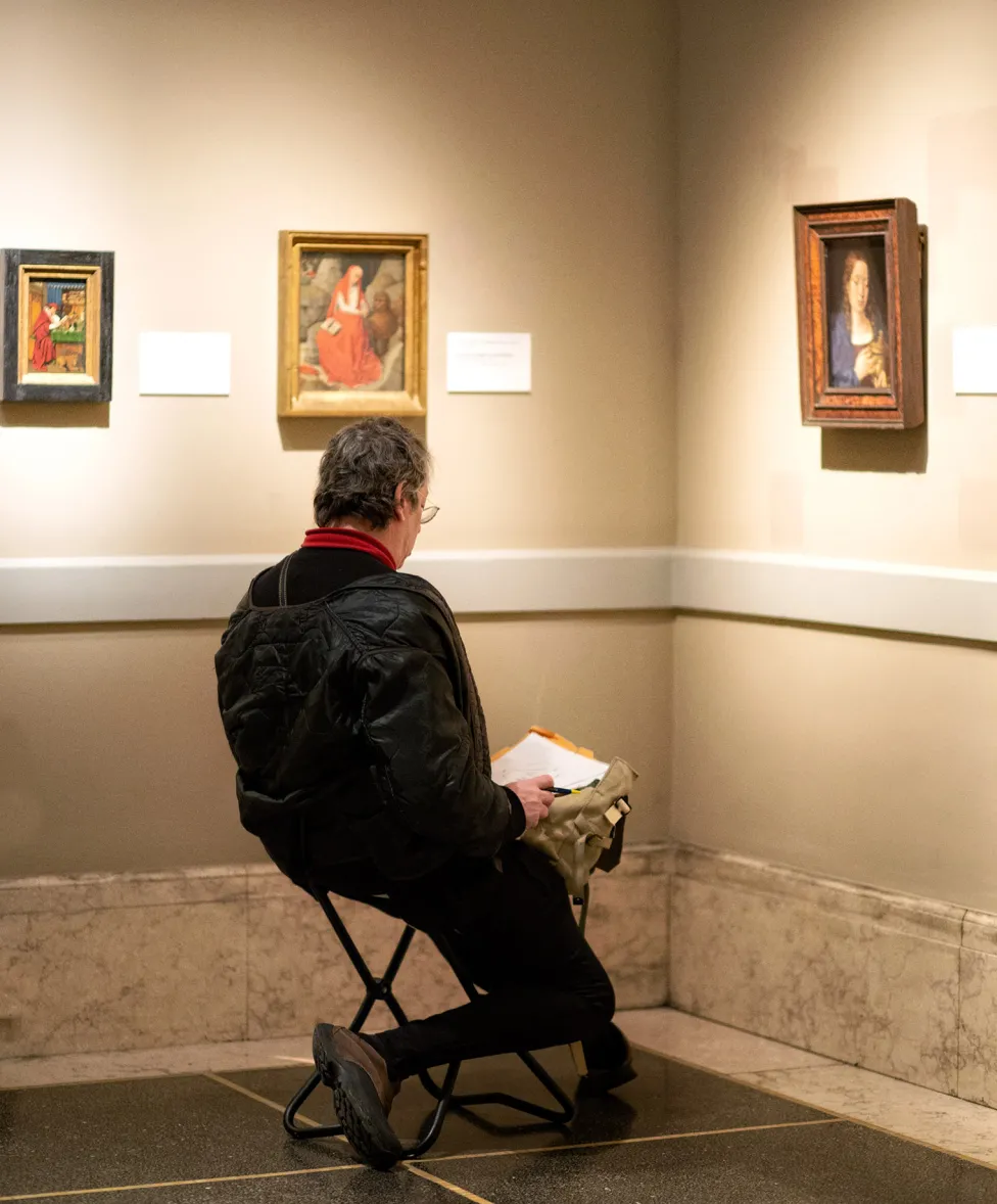 A visitor sitting on a stool in the European Galleries while drawing on their lap