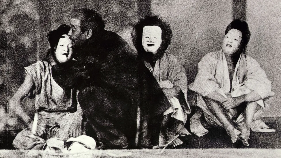 Three people sitting crossed legged on the ground in white masks with one being held by a man in black kneeling beside them.