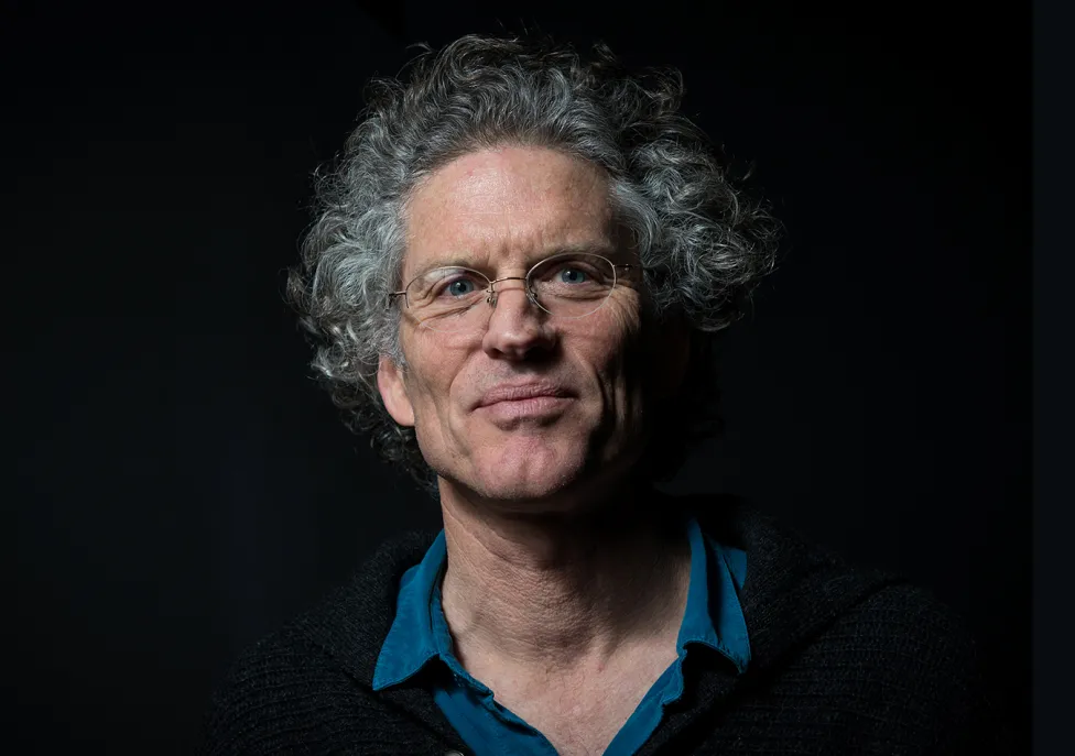 A headshot of poet David Hinton, wearing wire-rimmed glasses and a collared blue shirt, with curly gray hair.