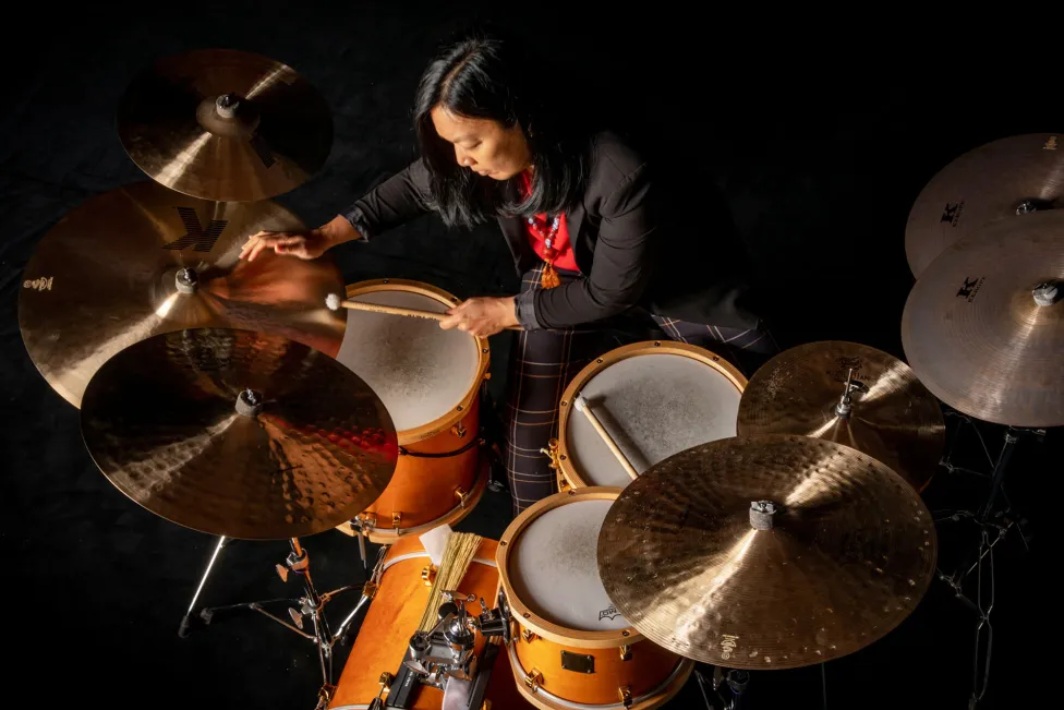 Susie Ibarra, wearing a suit with plaid pants, sits and plays a set of drums