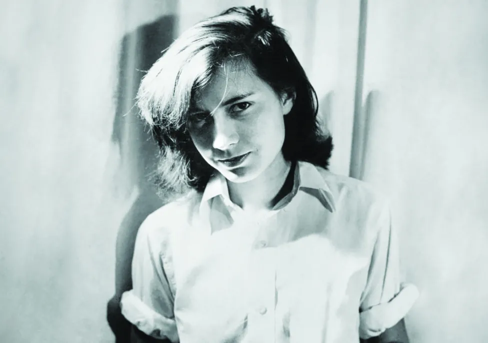 A young person pictured in black and white looks at the camera with tousled hair and a button down shirt with the sleeves rolled up past the elbows.