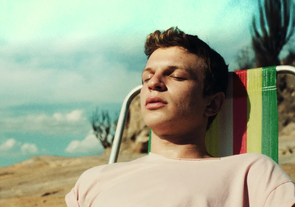 A young man rests on a colorful beach chair on a beach with his eyes closed.