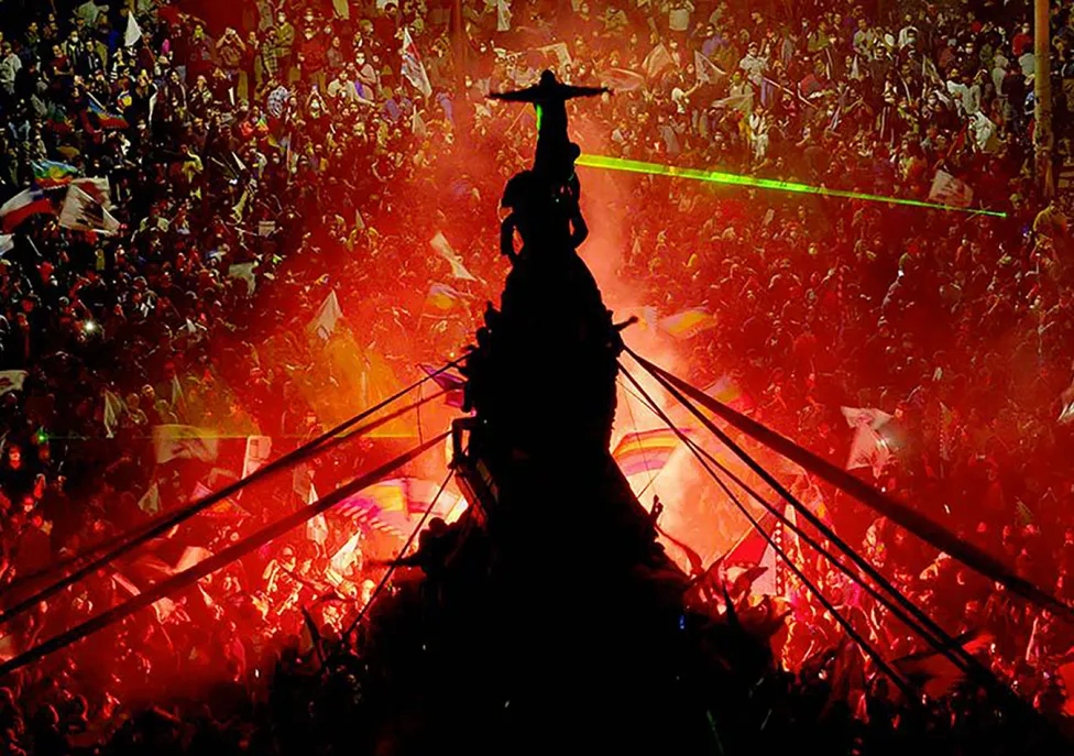 A thick crowd of people on the streets of Chile, with many standing atop a statue in the middle, while a red light emanates from the center.