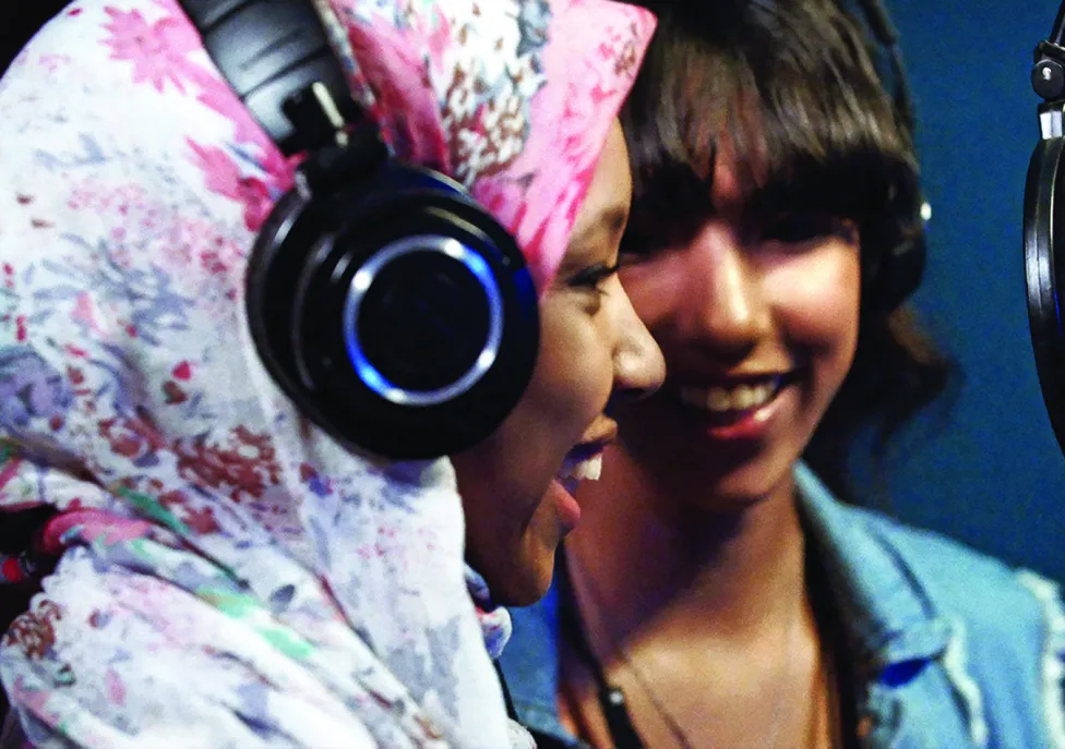 Two women wear over the ear headphones and laugh into a recording microphone