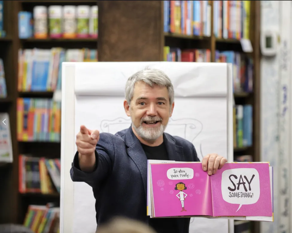 Author Peter Reynolds holding a book open to a bright pink spread and pointing to a crowd