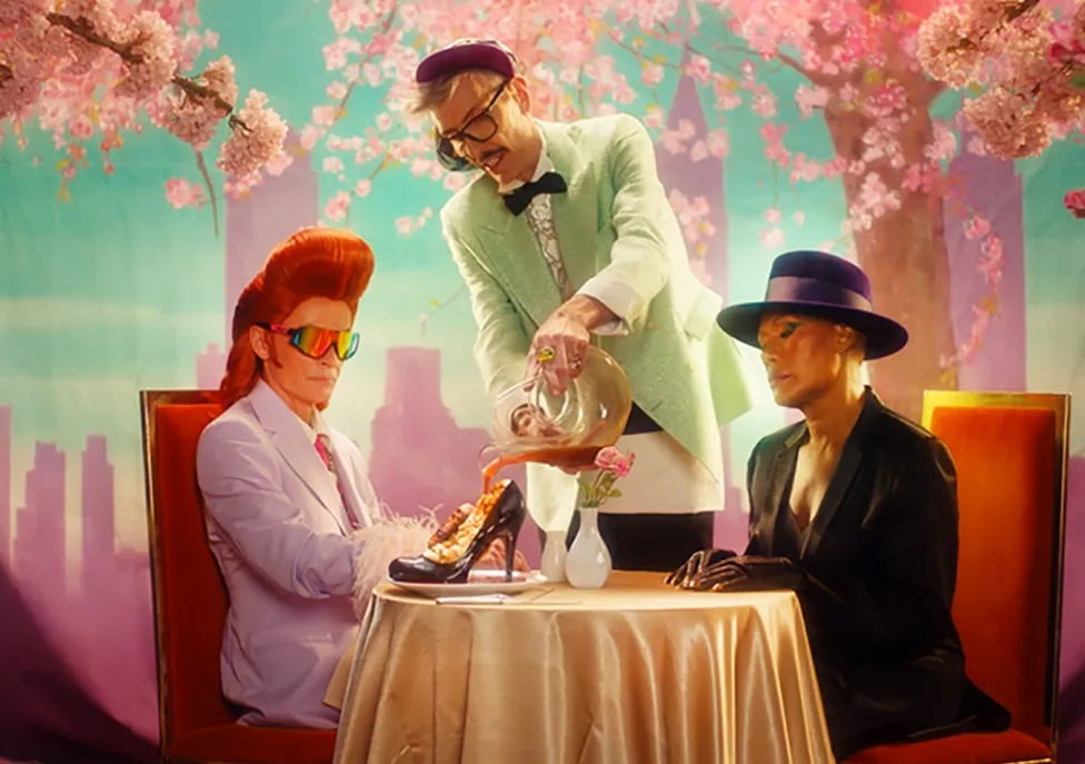 Two eclectic dressers sit at a table in front of a pink and teal hued skyline while a mint green clad waiter pours their caramel sauce into a high-heeled shoe.