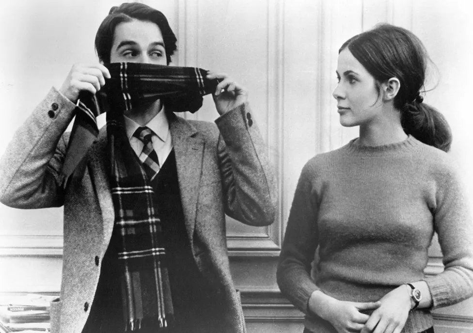 A man hides his lower face by holding up a plaid scarf that he's wearing while standing next to a woman who is looking at him.