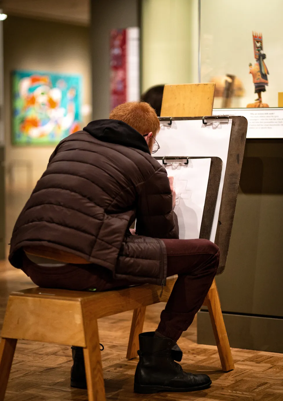 A visitor sitting on a drawing bench drawing on paper with a large clipboard in the Native American Galleries