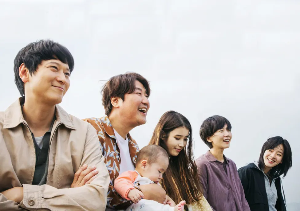 Two Korean men and three Korean women stand laughing in a row while one of the men holds a baby.
