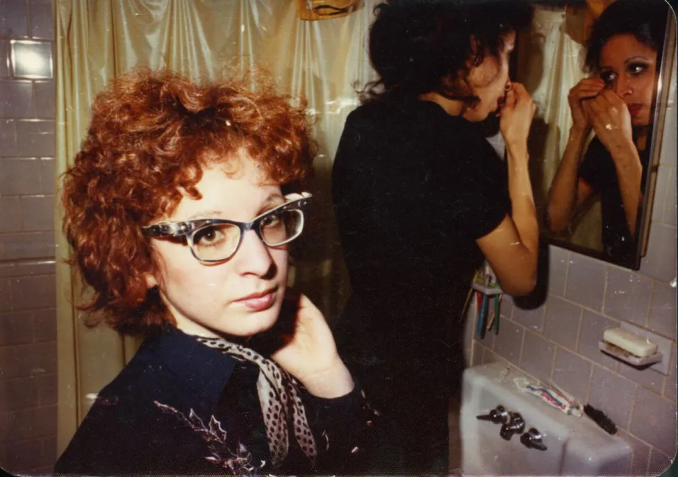 A woman with short, curly hair and glasses looks at the camera while another woman behind her looks closely at her face in a bathroom mirror.