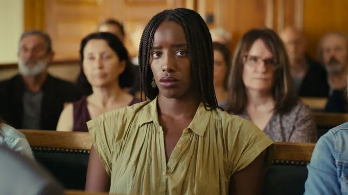 A Black woman wearing a collared shirt sits in a crowded courtroom gallery with a worried expression.