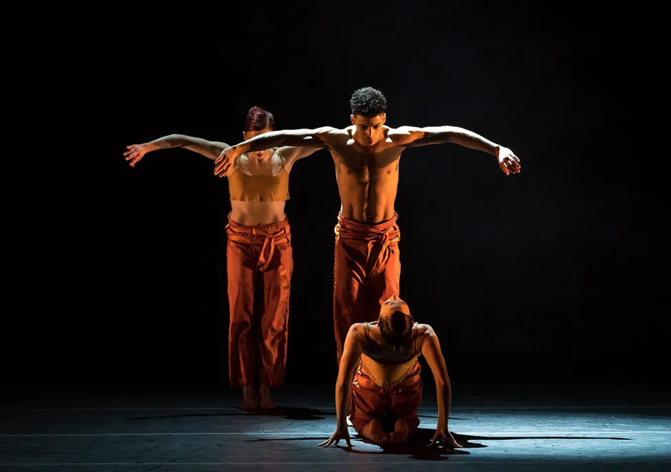 Three dancers on stage, two standing with their arms up and one sitting on the ground, looking backward towards the camera