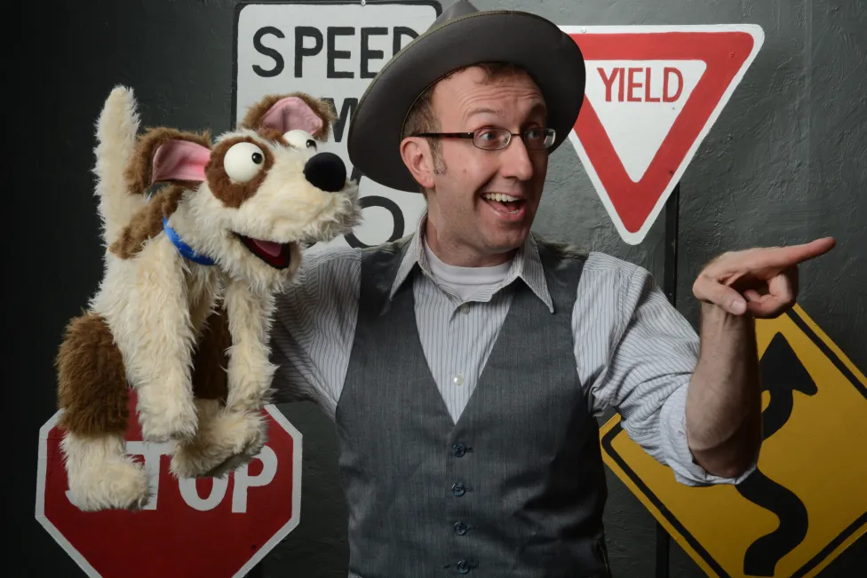 A man in a wide brimmed hat and vest holds a dog puppet in one hand and points out of view with the other. Various road signs are assembled behind him.
