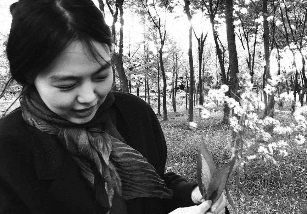 A woman bundled in a coat and scarf holds flowers picked in a forest, pictured behind her.