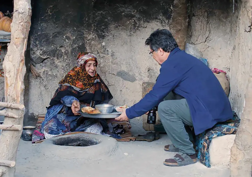 A man and a woman in a natural dwelling handing each other food over an in-ground fire pit