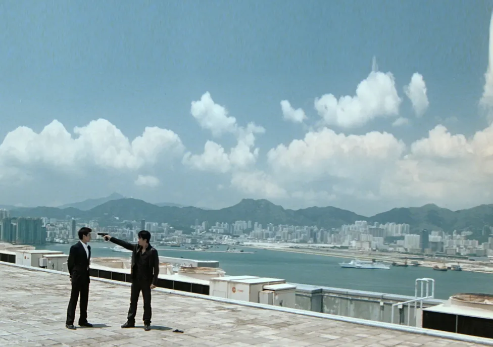 Two figures stand atop a high-rise roof with the city, a river, mountains and copious clouds behind them.