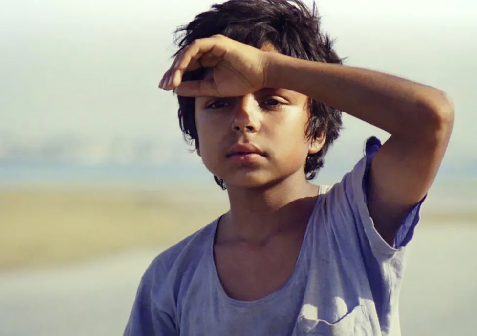 A young boy in a loose-neck t-shirt holds his hand up to his forehead as he looks out in the distance.