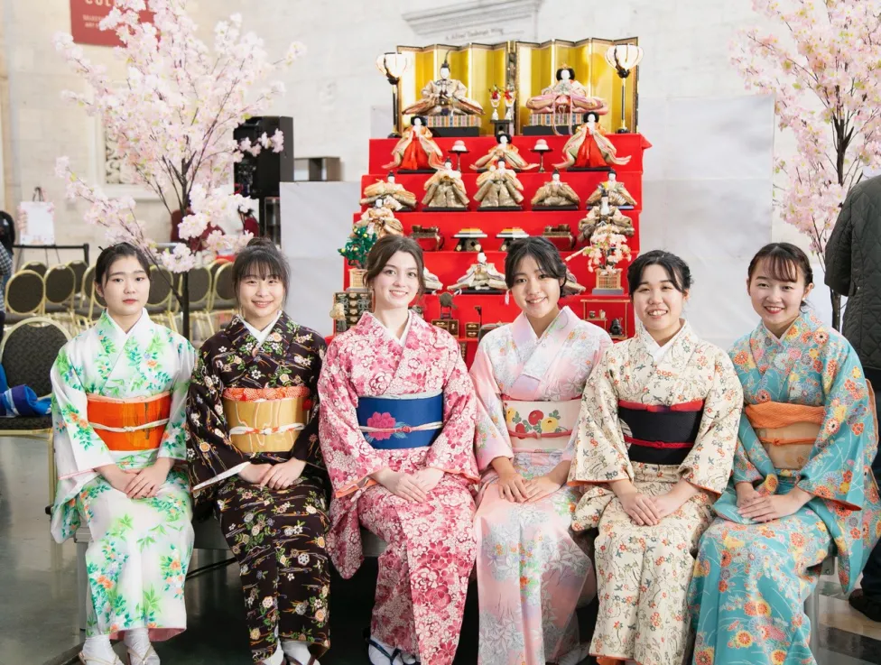 Six women, in traditional Japanese garments, sit in a row and smile at the camera.