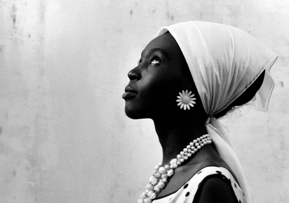 A black woman wearing a large flower earring and a scarf on her head looks up and to the left.