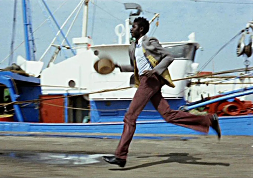 A man in a suit runs in front of docked boats while holding his hat.